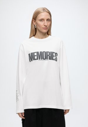 Žena s rovnými blond vlasmi má na sebe bielu košeľu s dlhým rukávom s čiernym textom "MEMORIES" a čiernu sukňu na jednoduchom svetlom pozadí.