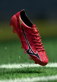 Rote Fußballschuhe mit geometrischem Muster, schwarzen und weißen Akzenten, Schnürverschluss und strukturiertem Obermaterial, über Gras dargestellt.