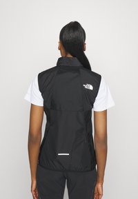 Svart ärmlös väst med hög krage, reflexdetaljer och logotyp på baksidan, tillverkad av lätt, vattentåligt material.