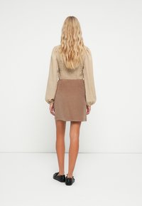 Blusa beige con fruncidos y mangas abullonadas, combinada con una falda corta de pana marrón y zapatos planos negros, vista desde atrás.
