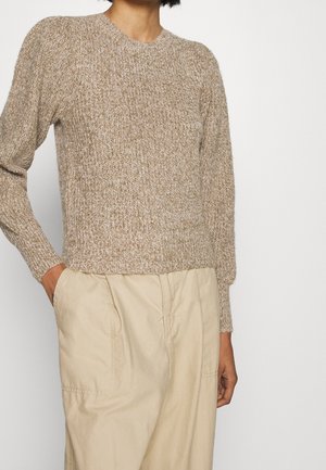 Beige gebreide trui met een ronde halslijn en geribbelde manchetten, met een gestructureerd patroon. Gecombineerd met lichtgekleurde, loszittende broeken.
