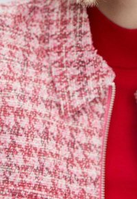 Gros plan sur une veste texturée rose et blanche avec une fermeture éclair, portée sur un haut rouge par une personne aux cheveux blonds.