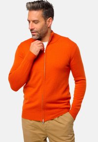 Cardigan orange zippé avec des poignets et un ourlet côtelés, fabriqué en tissu doux. Il présente un col haut et des détails de fermeture éclair lisses.