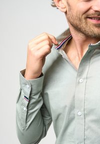 Heleda roheline nööbitega särk, millel on kaelus ja mis on kaunistatud punaste, valgete ja siniste triipudega. Pehme tekstuur, nähtav nööbiplokk ja reguleeritavad mansetid.