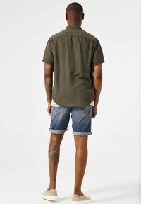 Korte mouwen, olijfgroene, knoopsluiting shirt gecombineerd met denimshorts. De shorts hebben omgeslagen zomen en achterzakken. Beigekleurige schoenen gedragen.