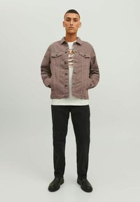 Giacca di jeans marrone chiaro con chiusura a bottoni e due tasche sul petto, indossata sopra una t-shirt bianca con grafica, abbinata a pantaloni neri e stivali.