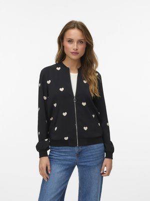 Hosszú hajú nő fekete, fehér szívekkel mintázott cipzáros dzsekit és kék farmert visel, tiszta világos háttér előtt áll.