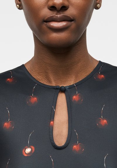 Zbliżenie osoby noszącej ciemną bluzkę z czerwonymi wzorami wiśni i dekoltem w kształcie otworu na klucz, zapinanym na pojedynczy guzik.
