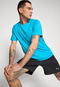 Lyseblå atletisk skjorte med korte ærmer, parret med sorte shorts. Funktioner inkluderer et Puma-logo på skjorten og tatoveringer på armene.
