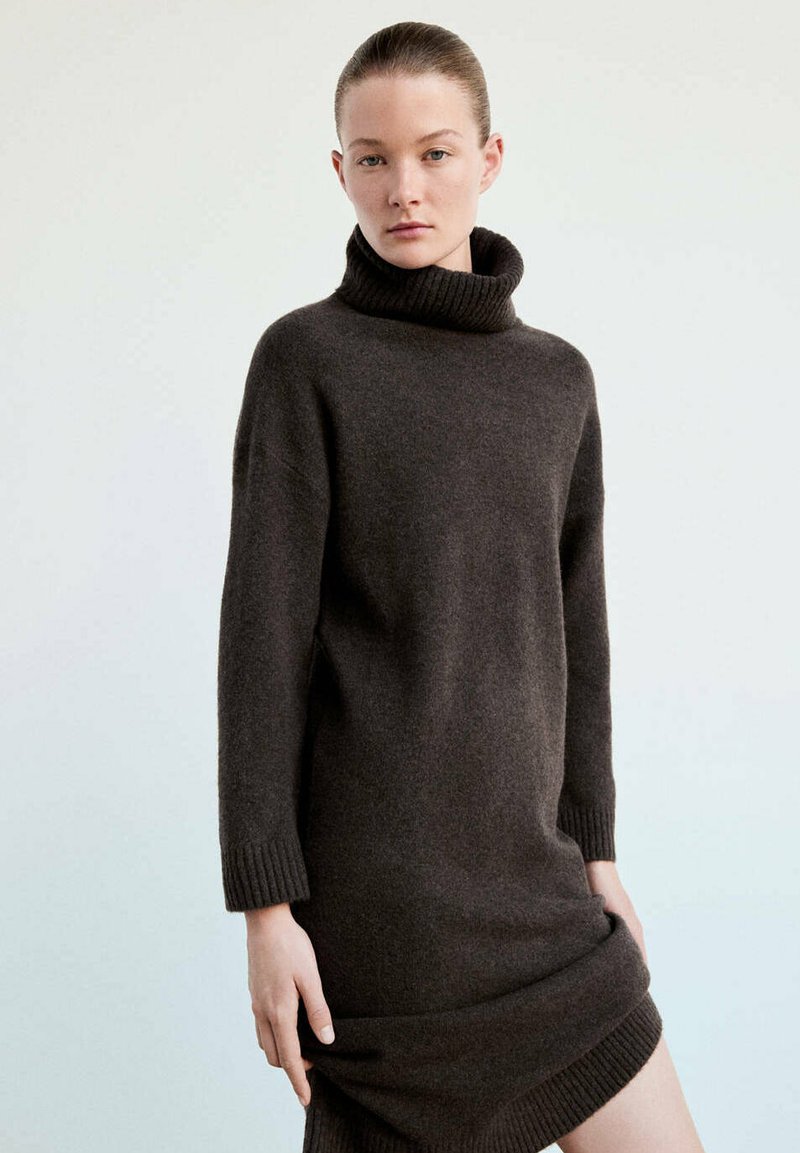 Jeune femme portant une robe-pull col roulé oversize marron foncé avec manches longues sur un fond clair uni.