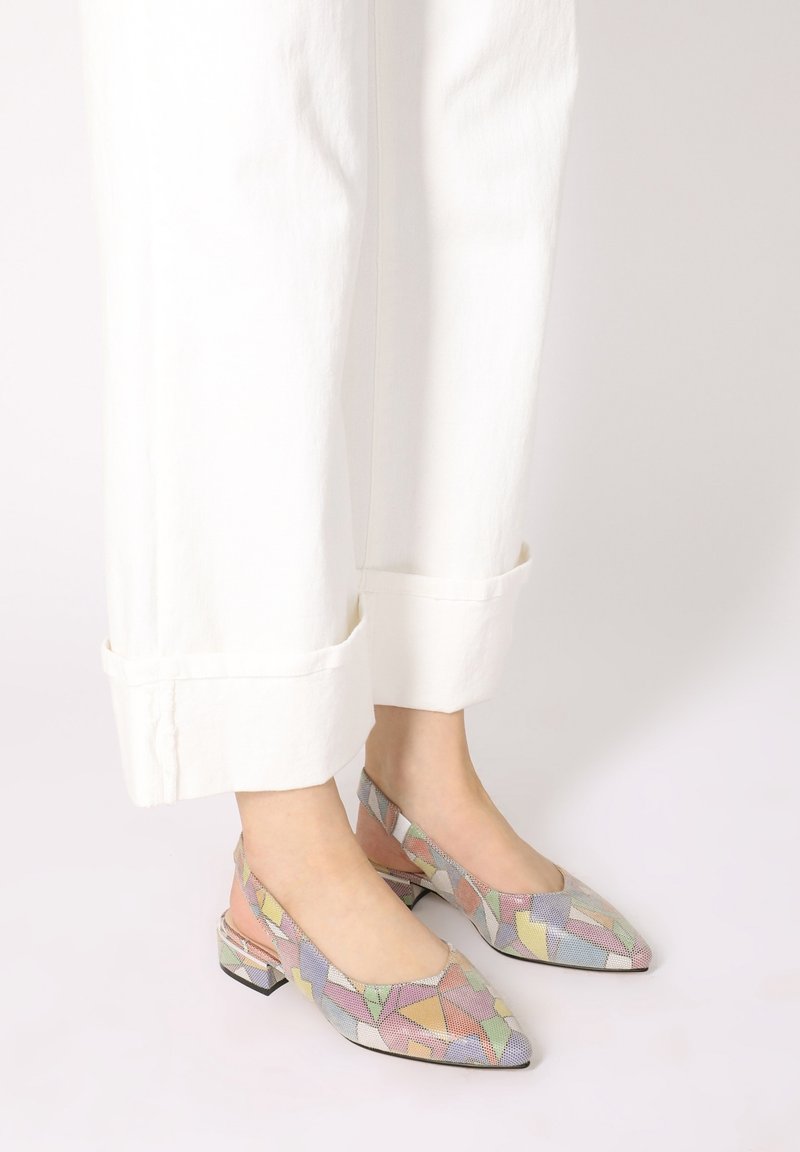Pieds portant un pantalon blanc retroussé et des chaussures slingback colorées à petit talon avec un motif géométrique, debout sur une surface claire.