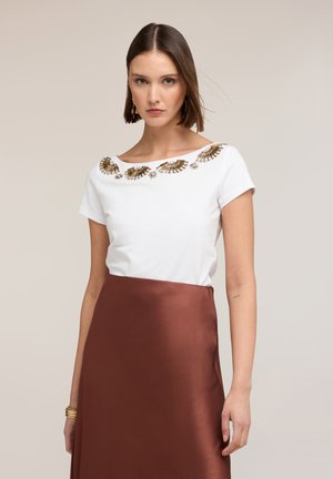 Frau trägt ein weißes kurzärmliges Oberteil mit goldenen, fächerförmigen Verzierungen und einen hoch taillierten braunen Satinsrock vor einem neutralen Hintergrund.