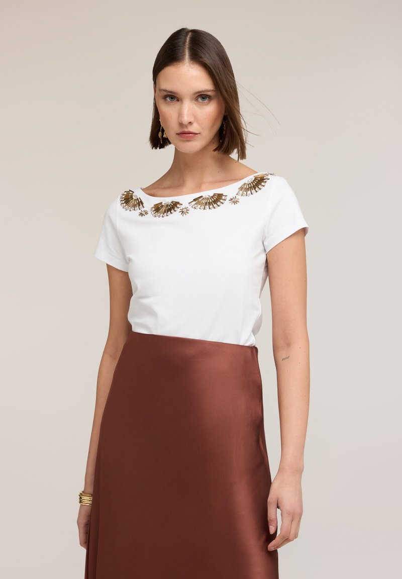 Kobieta ubrana w biały top z krótkim rękawem ozdobiony złotymi, wachlarzowatymi aplikacjami oraz brązową, satynową spódnicę z wysokim stanem na neutralnym tle.