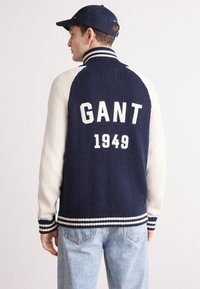 Námornícky a krémový hrubý pletený sveter s rebrovaným golierom, pruhovanými manžetami a veľkým potlačou "GANT 1949" na chrbte. Mäkčený, texturovaný povrch.