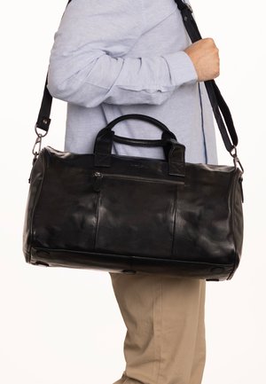 Persona con camisa azul claro y pantalones beige llevando una bolsa de viaje de cuero negro con asas y una correa ajustable para el hombro.