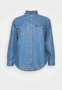 Camicia in denim azzurro chiaro con colletto classico, due tasche sul petto e orlo curvo. Il design include dettagli con bottoni in tutto.