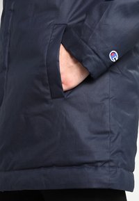 Marineblaue Außenjacke aus glattem Nylon, ausgestattet mit einer Seitentasche und einem Logopatch am Bündchen, im geraden Schnittdesign.