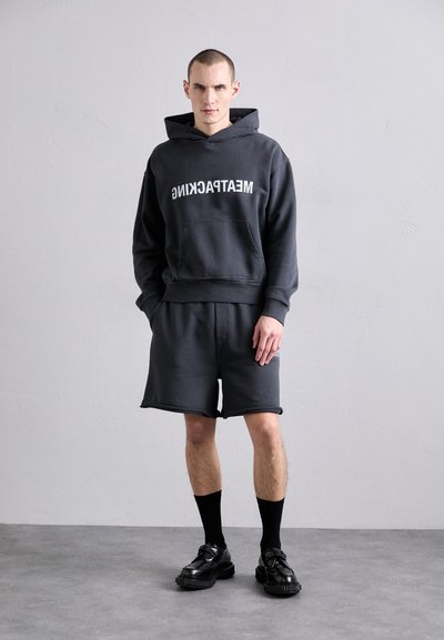 Mørkegrå hettegenser med frontlomme og fet hvit tekst, kombinert med matchende shorts. Myk, teksturert stoff, avsluttet med svarte sko og sokker.