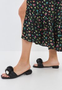 Sandali flat neri con design annodato e hardware decorativo. Texture in pelle liscia, stile open-toe e tacco basso a blocco.