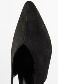 Svart mocka högklackad sko med spetsig tå och öppen sidsdesign. Slät yta med matt finish.