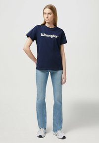Navy blue cotton t-shirt with "Wrangler" logo in cream, paired with light blue flared jeans and athletic shoes. Simple and casual design.