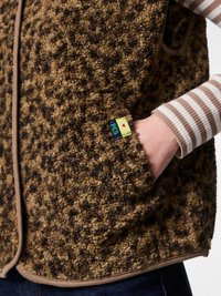 Close-up of a brown patterned fleece vest with a hand in the side pocket, worn over a beige and white striped long-sleeve shirt.