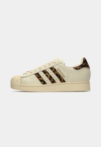 Off-white Adidas Superstar sneaker with leopard print stripes and heel tab, beige rubber shell toe, and "SUPERSTAR" text on the side.