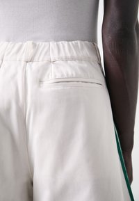 Vita byxor med en slät textur, elastisk midja och en enda bakficka. Har en grön accentremsa längs sidan.