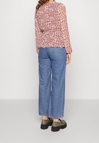 Donna che indossa jeans blu con orlo ripiegato, una blouse rossa a maniche lunghe con motivi floreali e mocassini verde con suola spessa, in piedi di spalle.
