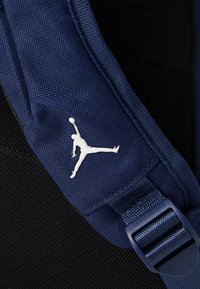 Sötétkék hátizsákpánt, texturált anyagból, állítható csattal és fehér Jumpman logóval. A háttér fekete �és hálószerű.