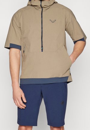 Homme portant une veste à capuche beige à manches courtes avec une fermeture éclair à l’avant et un short bleu marine, debout avec une main dans la poche devant un fond uni.