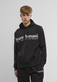 Hanorac cu glugă de culoare neagră, cu text alb "bruno banani. NOT FOR EVERYBODY" și o buzunar frontal, fabricat din țesătură moale cu manșete ribbate.