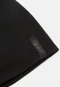 Mustat neulepipo, jossa on teksturoidumpi pinta ja musta logotagi. Piipon muoto on yksinkertainen ja pyöristetty, ja siinä on taitettu reuna.