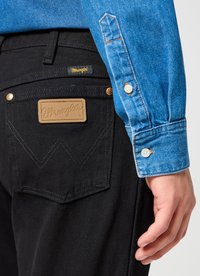 Person wearing black Wrangler jeans with a tan logo patch on the back pocket and a blue denim shirt with white buttons.
