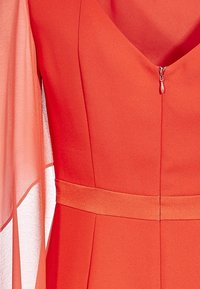 Robe rouge avec un col en V et une fermeture éclair dissimulée. Elle présente des manches longues en voile transparent et une taille cintrée avec une texture lisse.