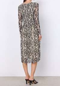 Maxikleid aus durchscheinendem, gemustertem Stoff mit langen Ärmeln und gerüschter Taille. Die Farben umfassen Beige, Braun und Grau. Schwarze Absatzsandalen.