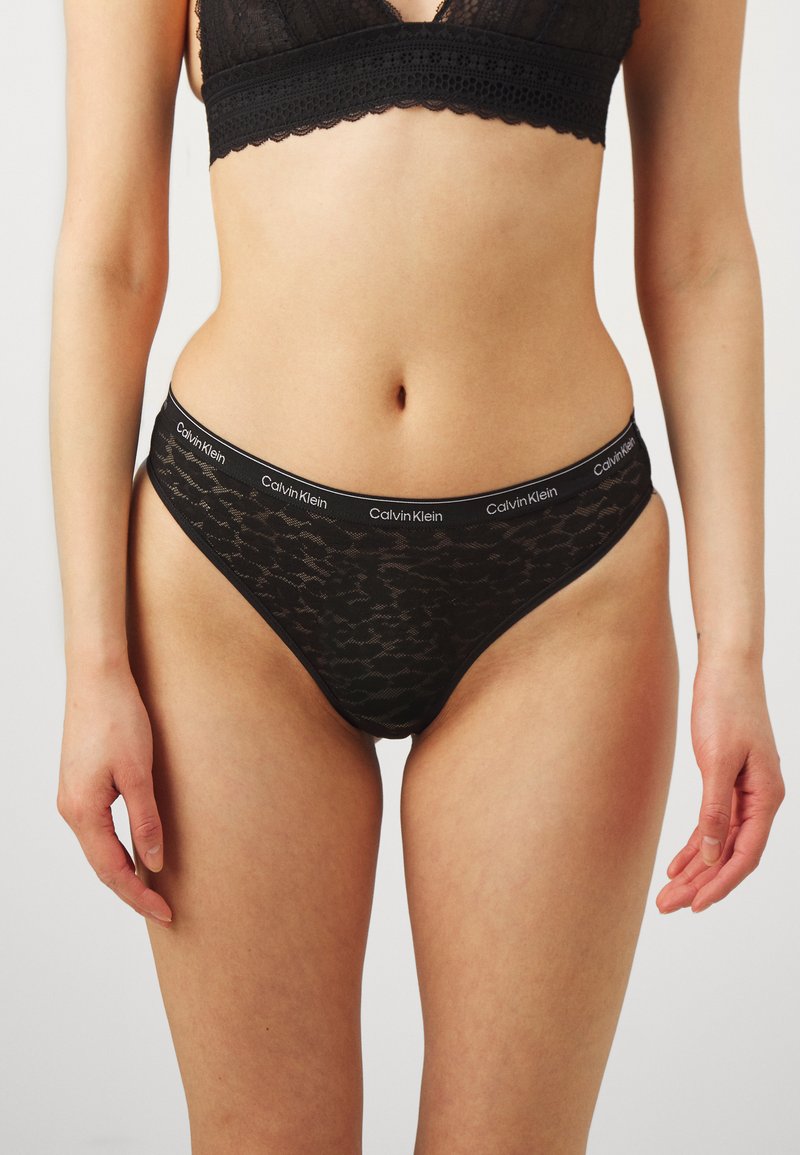 Tanga de encaje negro con un diseño estampado, que cuenta con una cinturilla suave con la marca "Calvin Klein". Textura suave, cobertura mínima.