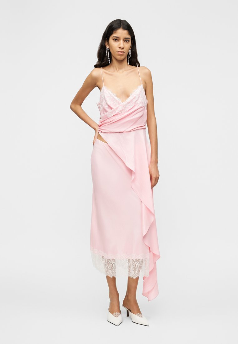 Robe rose à bretelles spaghetti avec des accents en dentelle, présentant un design drapé et un ourlet à volants. Modèle portant des chaussures blanches à bout pointu.