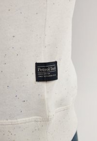 White sweatshirt with textured flecks in blue and green. Features a black label reading "Petrol Ind" and ribbed hem.