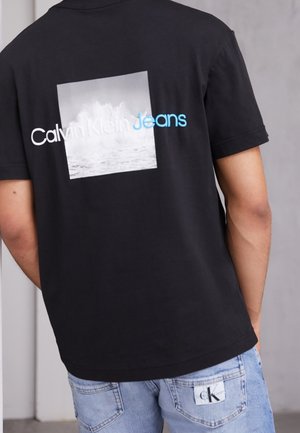 Sort bomulds T-shirt med en kvadratisk grafik af havbølger, med "Calvin Klein Jeans" i hvid og blå tekst på ryggen.