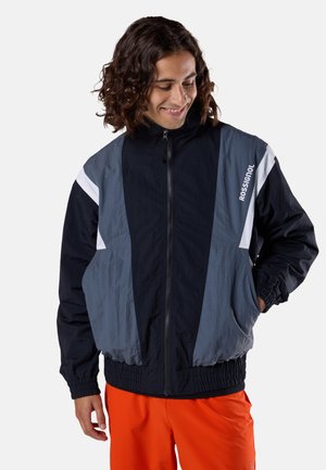 Nuori mies kiharatukkaisena, pukeutuneena musta-harmaaseen Rossignolin vetoketjutakkiin ja kirkkaan oransseihin housuihin, katsoo alas ja hymyilee.