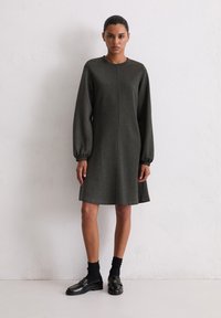 Robe A-line verte foncée en tissu doux, dotée de longues manches bouffantes et d'un col rond, associée à des mocassins noirs et des chaussettes.