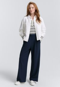Witte denimjas, gestreepte zwart-witte top, marineblauwe brede broek en witte sneakers. Model staat met handen in de zakken.