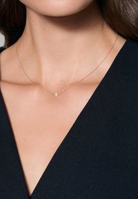 Złoty naszyjnik łańcuszkowy z delikatnym wzorem i małym akcentem w postaci perły na środku, na tle ciemnej tkaniny.