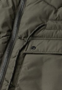 Detailní záběr na olivově zelenou péřovou bundu s viditelným zipem, šňůrkou a klopou kapsy na patentní knoflíky.
