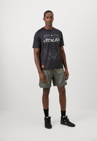 T-shirt de sport noir avec un design graphique blanc, associé à un short vert avec des motifs clairs. Porté avec des chaussures montantes noires et des chaussettes.
