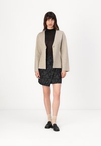 Beige pletený kardigan s výstřihem do V, dvěma knoflíky, kombinovaný s černou vzorovanou sukní, nošený přes černé dlouhé tričko.
