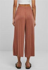 Pantalon large de couleur rouille en tissu doux. Il présente une taille élastique et des fentes latérales pour faciliter les mouvements.