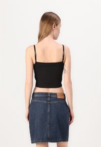 Svart avkortet topp med tynne stropper, kombinert med en blå denim miniskjørt. Skjørtet har et merkelapp på midjebåndet. Enkelt bakgrunn.