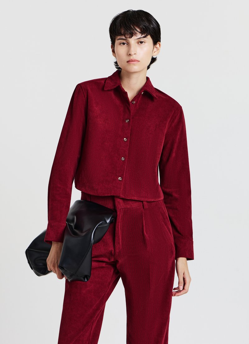 Camicia corta in velluto a coste rosso con colletto, bottoni frontali e maniche lunghe. Abbinata a pantaloni coordinati e con una borsa nera in mano.